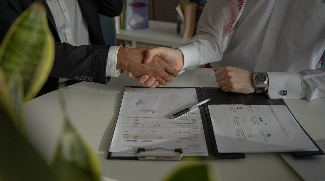 Two people shaking hands over signed documents at a table.